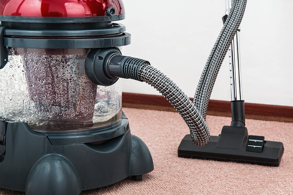 A person wearing beige shoes and blue jeans is operating a black and silver vacuum cleaner with a yellow canister on a patterned, traditional-style area rug in a room. The rug features a floral design
