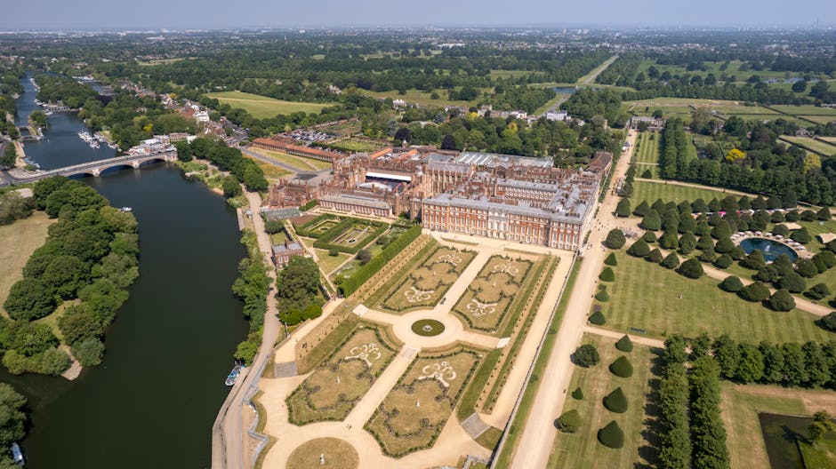 Aerial view of Hampton Court Palace in Surrey showcasing its historic brick and stone façade, surrounded by lush green gardens, neatly trimmed lawns, and tree-lined pathways. The palace is positioned