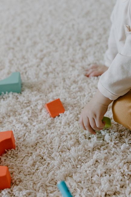 Close-up of a child's hand resting on a plush, cream-colored carpet with a high pile, situated in a well-lit living room or play area. The carpet appears clean and soft, with a few colorful toy blocks